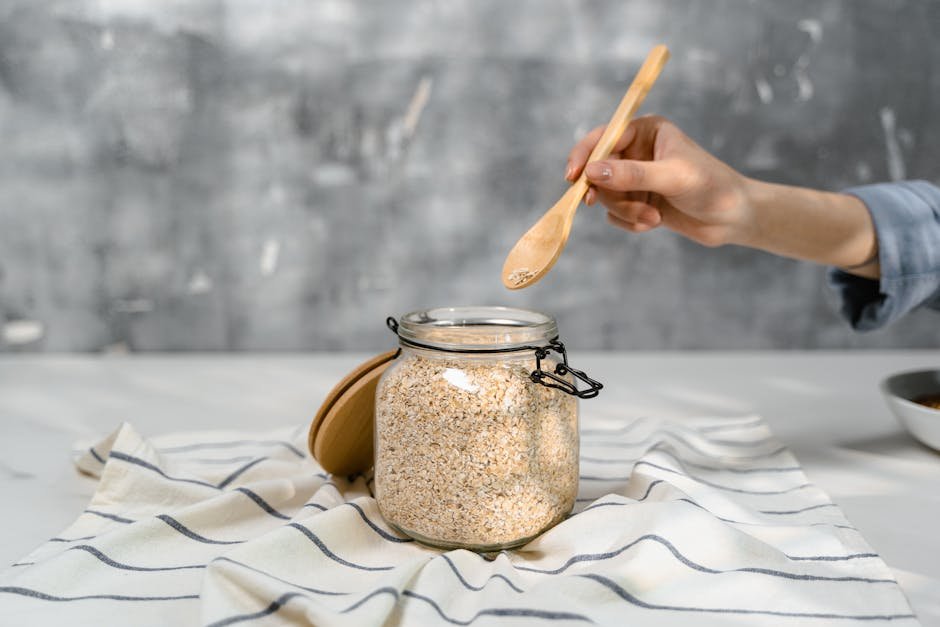 Hand holding a wooden spoon over a glass jar filled with oats on a striped tablecloth.